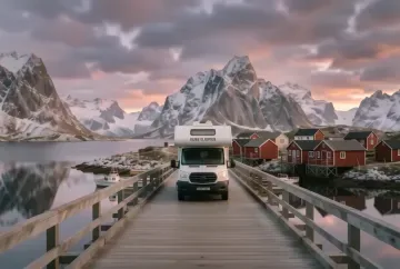 Wohnmobil auf einer Lofoten-Brücke nahe Reine mit roten Rorbu-Hütten am Wasser und Bergen im Sonnenaufgang