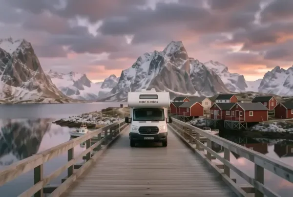 Wohnmobil auf einer Lofoten-Brücke nahe Reine mit roten Rorbu-Hütten am Wasser und Bergen im Sonnenaufgang